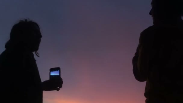 Siluety muže a ženy s jejich chytré telefony proti barevné nebe - Záběry, video