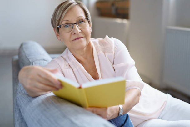 Senior woman sitting on sofa with book - Foto, afbeelding