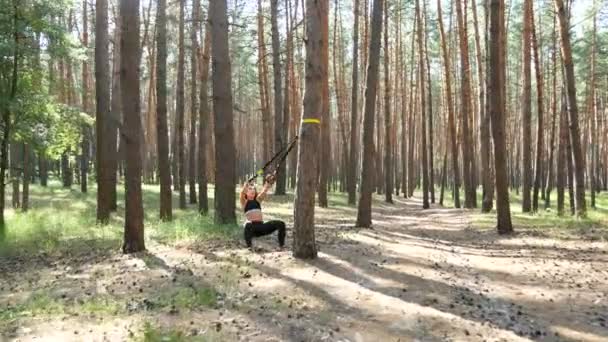 Beautiful, athletic, sexy young woman, coach, instructor, performs exercises, doing exercises with fitness trx system, TRX suspension straps. In pine forest, in summer, in sun rays. - Video, Çekim