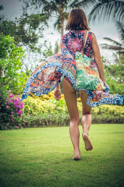 Belle jeune femme en paréo et maillot de bain marchant dans le parc becah avec sac le jour ensoleillé d'été. Tropical île de Bali, Indonésie
. - Photo, image