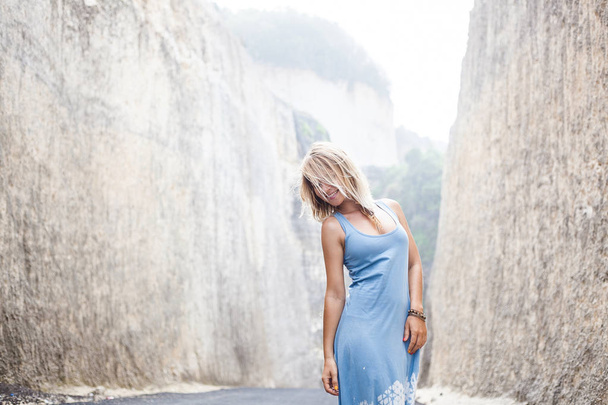 Horizontal portrait of an attractive smiling girl in light-blue dress is standing on the road between mountains and looking away - Foto, Bild