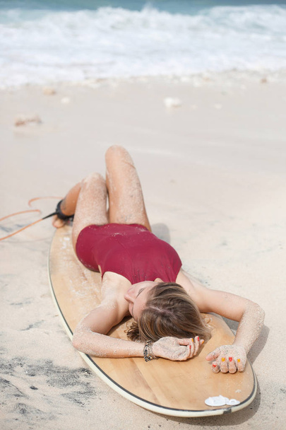 Attractive surf girl with closed eyes in dark-red swimming suit is sprawling on a surfboard on the beach, reaching out her hands - Valokuva, kuva