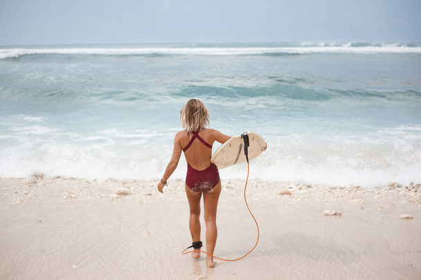 Retrato panorâmico horizontal de uma menina de surf atraente em terno de natação vermelho escuro está indo para o oceano com uma prancha de surf, vista de trás
 - Foto, Imagem