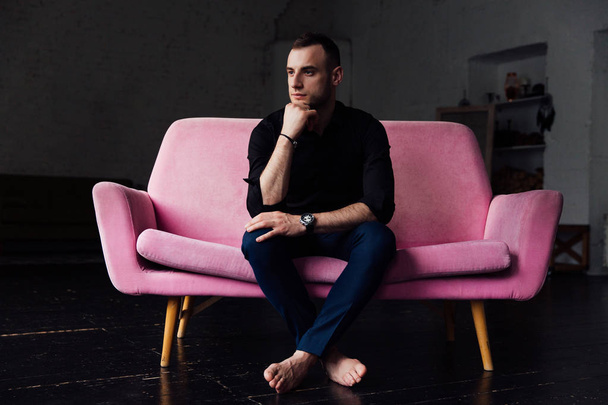 Man model sits on pink sofa in loft interior. Businessman in a black shirt and watch, blue pants. - Foto, afbeelding