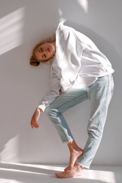 Uma menina bonita e doce em jeans e uma camisola fica perto de uma parede branca
 - Foto, Imagem