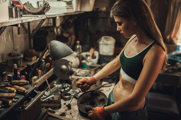 Εργαζόμενο κορίτσι σε t-shirt κάθεται στο εργαστήριο μεταξύ εργαλείων και ξεβιδώνει - Φωτογραφία, εικόνα