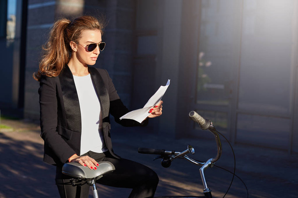 Environmentally friendly female manager in formal suit and sunglasses reading report with serious face expression. Stylish and attractive Caucasian woman stops her bicycle on the way to the office - Photo, Image
