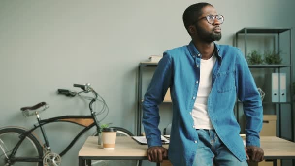 Portrait of young attractive african business man with glasses in denim blue shirt looking on camera and smiling in the office background while leaning on the office table. - Metraje, vídeo