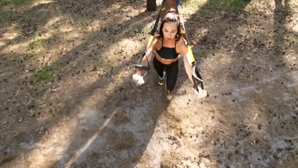 Beautiful, athletic young woman, coach, instructor, performs, doing exercises with fitness trx system, TRX suspension straps. In pine forest, in summer, in sun rays. view from above - Imágenes, Vídeo