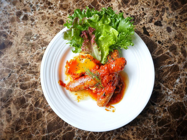 вид на вкусные копченые колбасы в томатном соусе с овощами на белой тарелке на мраморном столе
 - Фото, изображение
