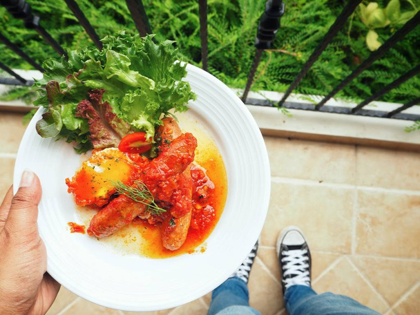 Вид сверху на человека, держащего тарелку с вкусными копчеными сосисками в томатном соусе
 - Фото, изображение