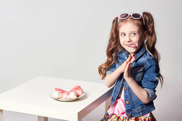 Fashion fun red-haired girl with pigtails and goggles on her head wants to eat a cake, a happy facial expression. Studio photo on light coloured background. Birthday, holiday - Fotó, kép