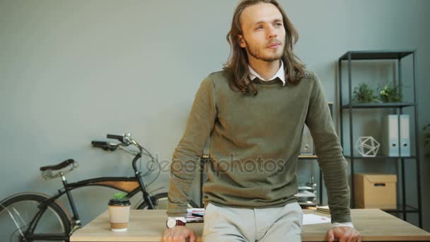 Young caucasian man with long hair crossing hands, looking to the camera and smiling in the office background while leaning on the office table. - Filmmaterial, Video