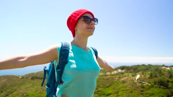 Una mujer con una mochila detrás de su espalda gira sus brazos hacia los lados, de pie en la cima de una colina en la orilla del océano
 - Imágenes, Vídeo
