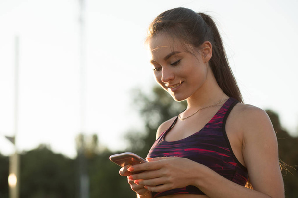 Sportswoman using smart phone outdoors. Female athlete in sportswear on stadium outdoors. Modern technology concept, sport activity. - 写真・画像