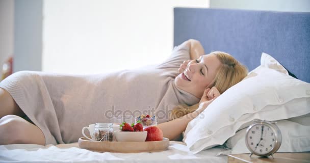 Beautiful woman eating breakfast with yogurt, fruit and cereals on bed. Morning wake up at home in bedroom. Caucasian people liying on bed. 4k video - Footage, Video