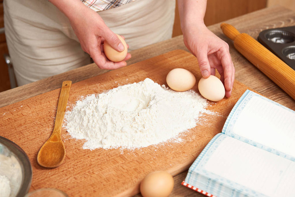 Vrouw koken deeg. Meel voor het bakken op een houten achtergrond met keukengerei. Handen en voedsel close-up. Een ei breken. - Foto, afbeelding