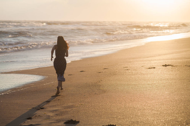 Tyttö mekko kulkee hiekkaranta pitkin myrskyinen meri auringonlaskun aikaan
 - Valokuva, kuva