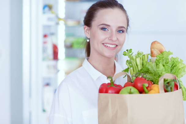 Young woman holding grocery shopping bag with vegetables. Standing in the kitchen. - Foto, imagen