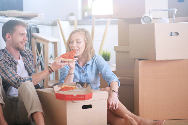 Young couple have a pizza lunch break on the floor after moving into a new home with boxes around them. Young couple - Zdjęcie, obraz
