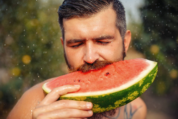 Bir adam şık bir saç modeli, uzun sakallı ve büyük bir zevk ile dövme karpuz yiyor - Fotoğraf, Görsel