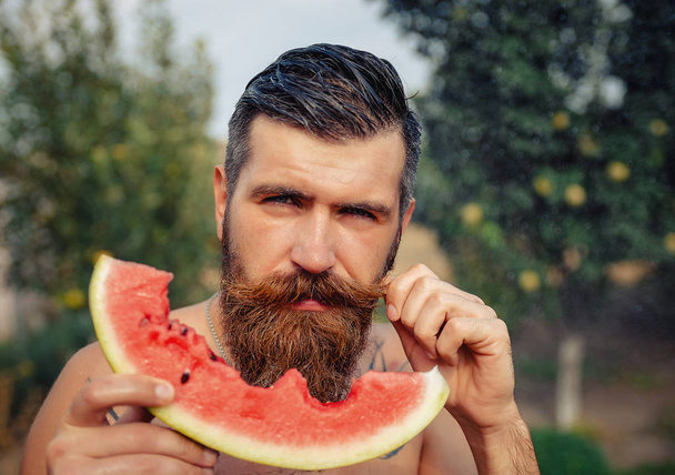 Um homem com um penteado elegante, uma longa barba e uma tatuagem com grande prazer come melancia
 - Foto, Imagem