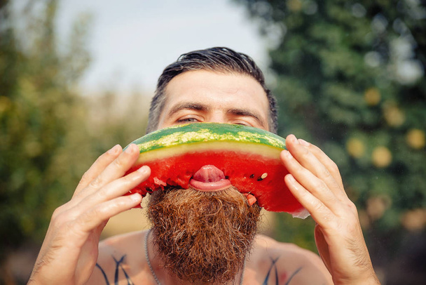A man with a stylish hairstyle, a long beard and a tattoo with great pleasure eats watermelon - Photo, Image