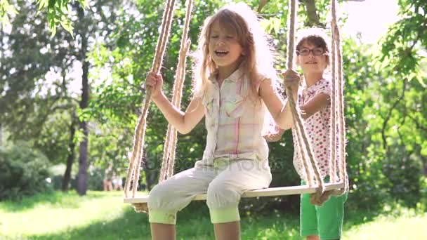 kinderen swingen op een schommel in de zomer Park. twee zusjes spelen in de frisse lucht. de oudere zus schudt haar jongere zus op de schommel. Swing op kabels aangesloten op de boom - Video