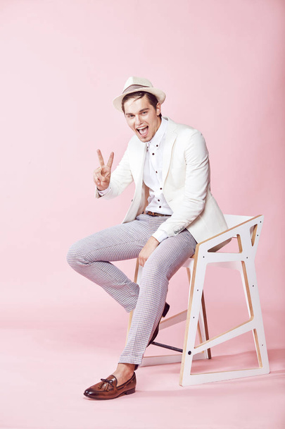 Young smiling man wearing white jacket, white shirt, grey pants and white hat sitting on the chair in studio with light pink background - Fotó, kép