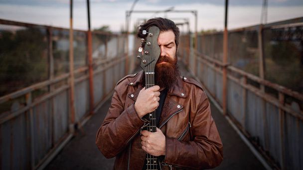 Stylishly dressed handsome man with a long beard, brown leather jacket and light jeans holding a guitar and standing on a bridge - Photo, Image