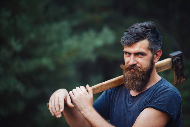 Elegante hombre barbudo con una camiseta oscura, con una cabeza en la cabeza sosteniendo en sus manos un hacha de madera en el bosque - Foto, Imagen