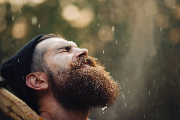 Siyah tişörtlü şık sakallı adam, kafasında kafasıyla ormanda tahta bir balta tutuyor. - Fotoğraf, Görsel