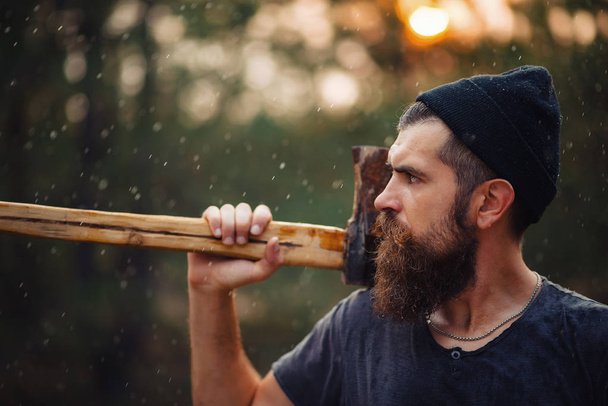 Stylish bearded man in a dark T-shirt, with a head on his head holding in his hands a wooden ax in the woods - 写真・画像