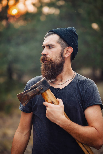 Стильный дровосек с длинной бородой и в модной одежде курит сигарету и держит топор в руках в лесу.
 - Фото, изображение