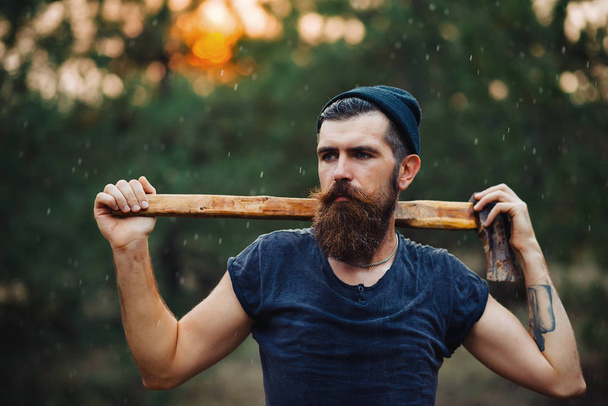 Stylowy brodaty mężczyzna w ciemnej koszulce, z głową na głowie trzymającą w dłoniach drewnianą siekierę w lesie - Zdjęcie, obraz