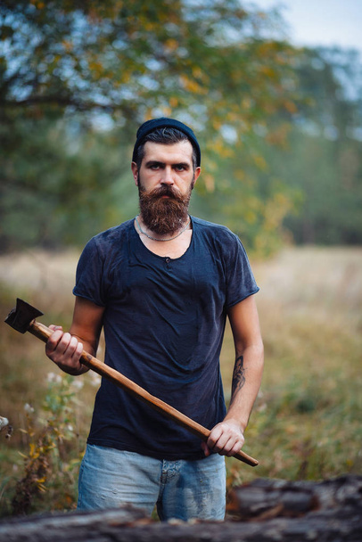 Стильний бородатий чоловік у темній футболці, з головою на голові тримає в руках дерев'яну сокиру в лісі
 - Фото, зображення