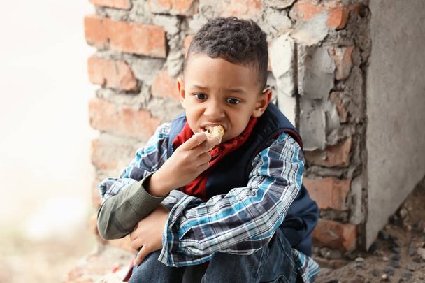 Cute little boy sitting on floor with piece of bread in abandoned building. Poverty concept - 写真・画像