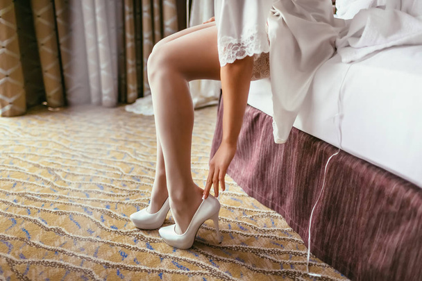 Невеста в шелковом халате с красивыми стройными ногами надевает свадебные туфли. Подготовка к свадьбе
 - Фото, изображение