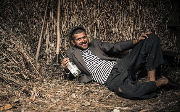 Een vrolijke boertje in oude kleren met een fles alcohol in zijn handen legt op een droog gras in een schuur - Foto, afbeelding