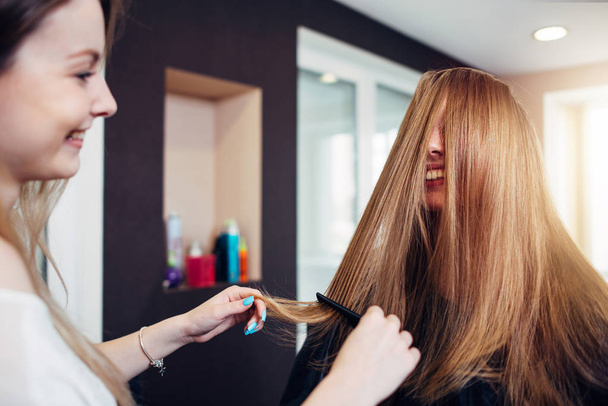 Cliente femenina con el pelo largo y liso riéndose mientras una peluquera peina hebras delante de su cara trabajando en el salón de belleza
. - Foto, Imagen
