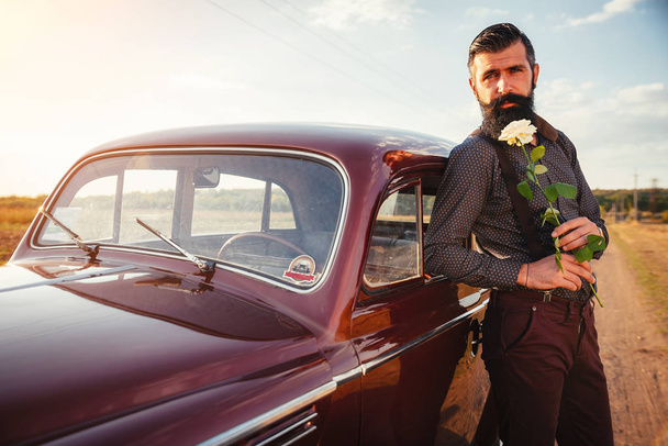 Elegantní vousatý muž s bílou růži čeká na své milované poblíž jeho červené auto - Fotografie, Obrázek