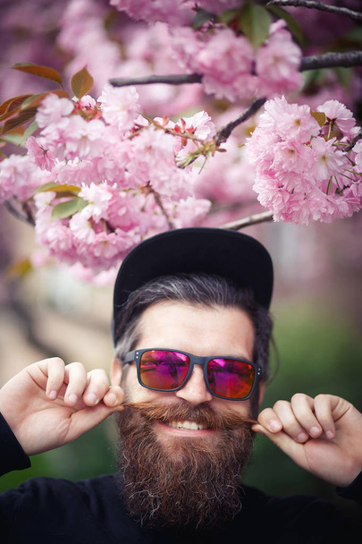Brutální vousatý muž s kloboukem a sluneční brýle, stojící poblíž kvetoucí sakura - Fotografie, Obrázek