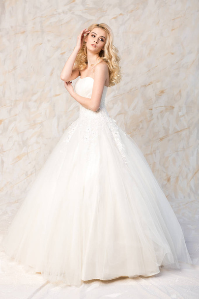 fashionable gown, beautiful blonde model, bride hairstyle and makeup concept - young lovely girl in white wedding festive dress, standing indoors on light background, romantic slender woman posing. - 写真・画像
