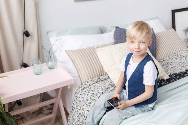 Rapaz pequeno inteligente com telefone móvel, assentos na cama no quarto. Conceito de tecnologia
 - Foto, Imagem