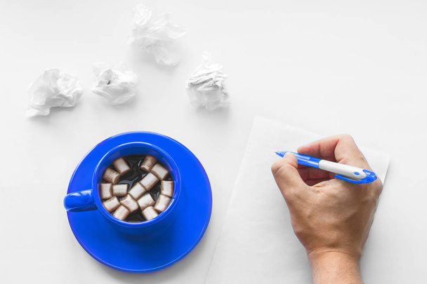 Cup of coffee on saucer with marshmallows, hand with pen writing on a blank sheet of paper on the wooden white background and crumpled sheets of paper. Business plan. Writing poetry. Never give up. - Фото, зображення