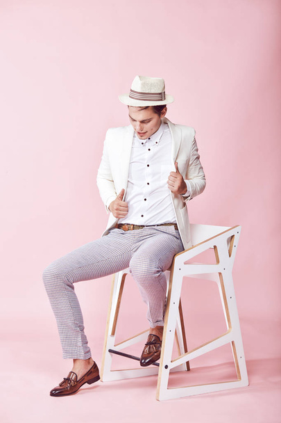 Young beautiful modern male model wearing white jacket, white shirt, grey pants and white hat sitting on the chair in studio with light pink background - Fotografie, Obrázek