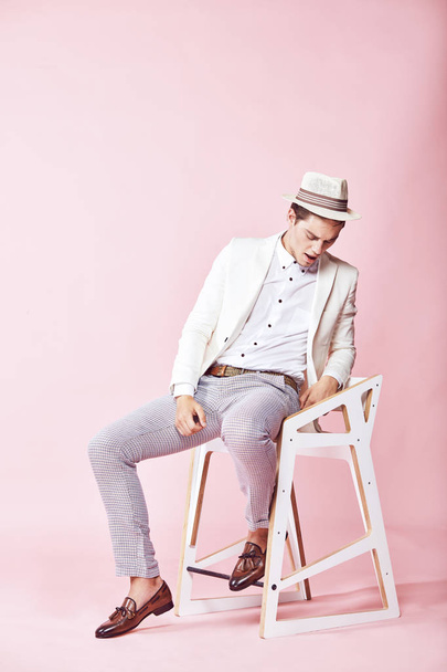 Young handsome happy modern male model wearing white jacket, white shirt, grey pants and white hat sitting on the chair in studio with light pink background - Fotografie, Obrázek