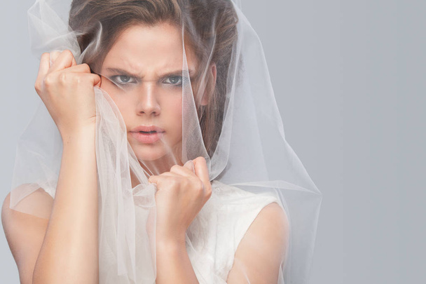 Angry bride with fashion wedding hairstyle - on gray background. Closeup portrait of young gorgeous bride. Wedding. Studio shot with copy space. Beautiful bride portrait with veil over her face - Photo, image
