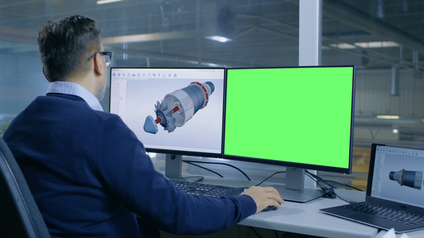 Главный инженер проектирует 3D турбину / двигатель для крупной промышленной компании, его второй дисплей показывает макет зеленого экрана компьютера. Из окна офиса видели большую фабрику
. - Кадры, видео