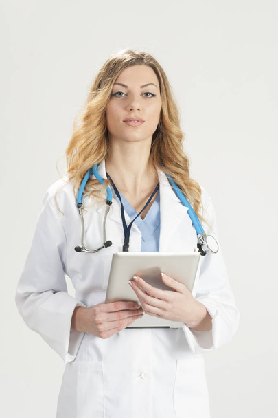 Женщина-врач в белом хирургическом халате со стетоскопом и таблеткой в руках, стоящих изолированно на белом фоне
 - Фото, изображение